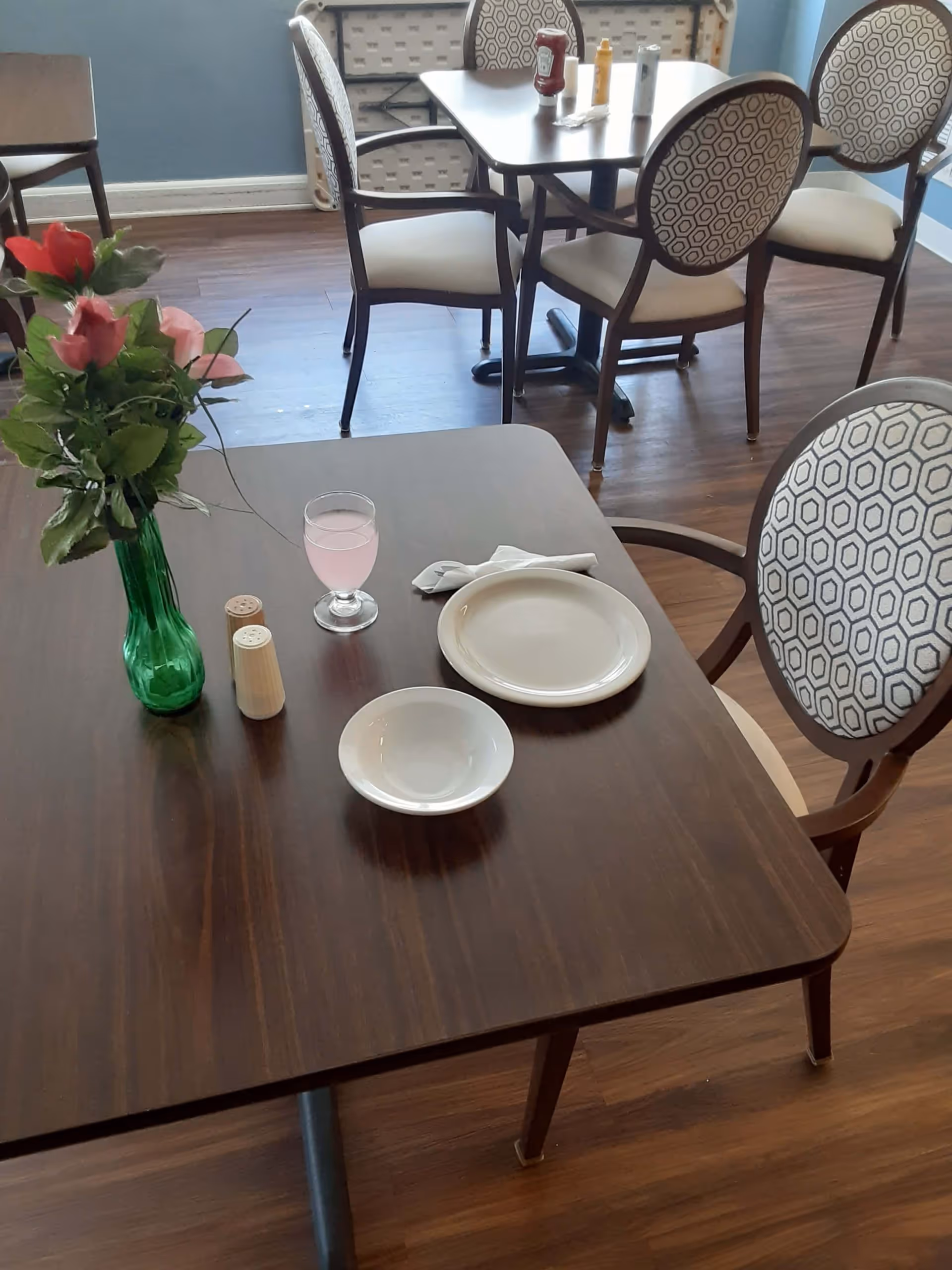 A dining table set with a plate, bowl, napkin, glass of pink beverage, salt and pepper shakers, and a green vase with red and pink flowers. In the background, there are more tables and chairs with patterned upholstery on the backrests, all set on a wooden floor.