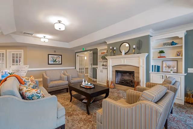 A cozy living room in a senior living facility featuring a fireplace with a white mantel, built-in shelves with decorative items, a dark wooden coffee table, and several upholstered armchairs and sofas arranged around the table. The walls are painted in neutral tones with a green accent wall around the fireplace, and the room is well-lit with ceiling lights.