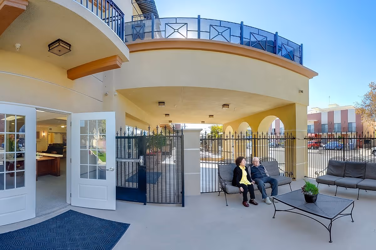 Outdoor covered patio area at Silverado Beverly Place Memory Care Community with two elderly people sitting on cushioned metal benches. The patio has a black metal fence, a table with a potted plant, and open double doors leading inside. The sky is clear and blue.