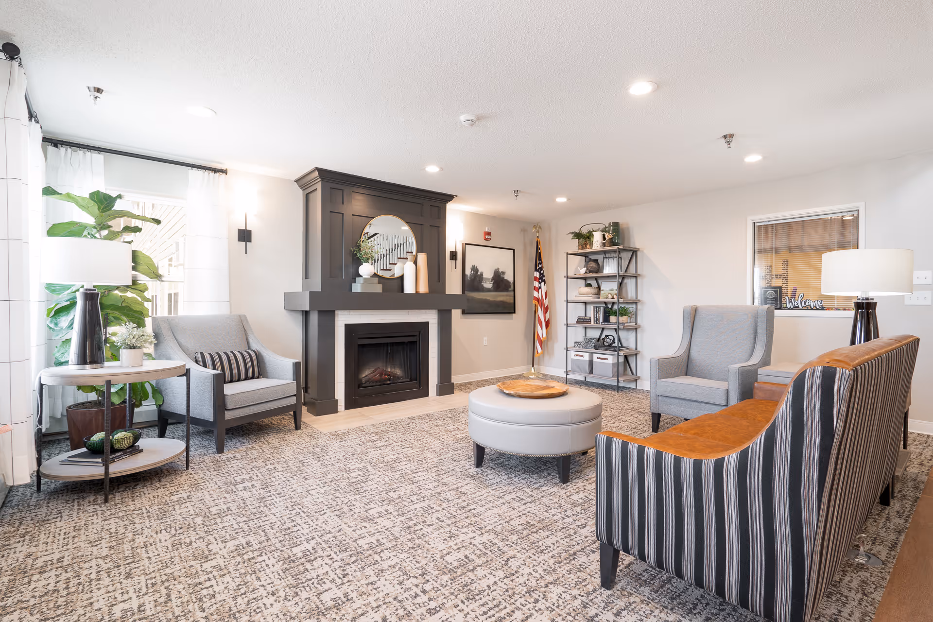 A bright and cozy living room area with a dark gray fireplace as the focal point. The room features a striped sofa with a brown leather back, two gray armchairs, a round gray ottoman, and a side table with a lamp and plants. There is a large window with white curtains letting in natural light, a bookshelf with decorative items, and an American flag in the corner. The walls are light-colored, and the floor is covered with a patterned carpet.
