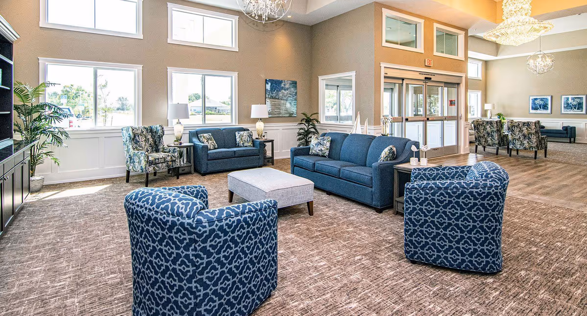 A spacious and well-lit living room area with multiple blue patterned armchairs, two blue sofas, a gray ottoman, side tables with lamps, and large windows letting in natural light. The room has beige walls, carpeted flooring, decorative plants, and modern chandeliers hanging from the ceiling. There is a glass entrance door visible in the background.