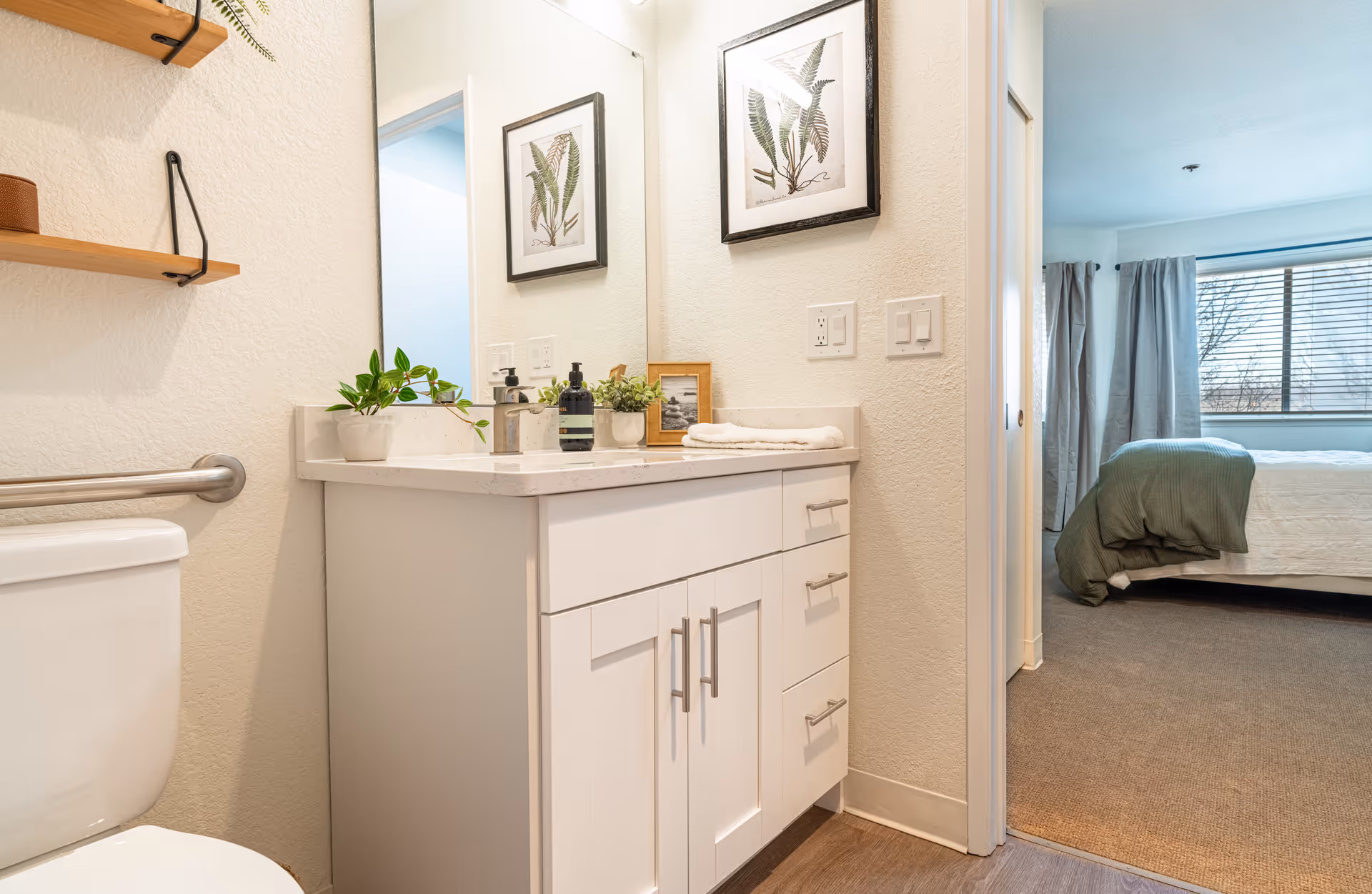 A bright bathroom with a white vanity cabinet topped with a marble countertop, a sink, and decorative plants. A toilet is partially visible on the left. The bathroom opens into a bedroom with a bed covered in white and green bedding, a window with blinds, and gray curtains.