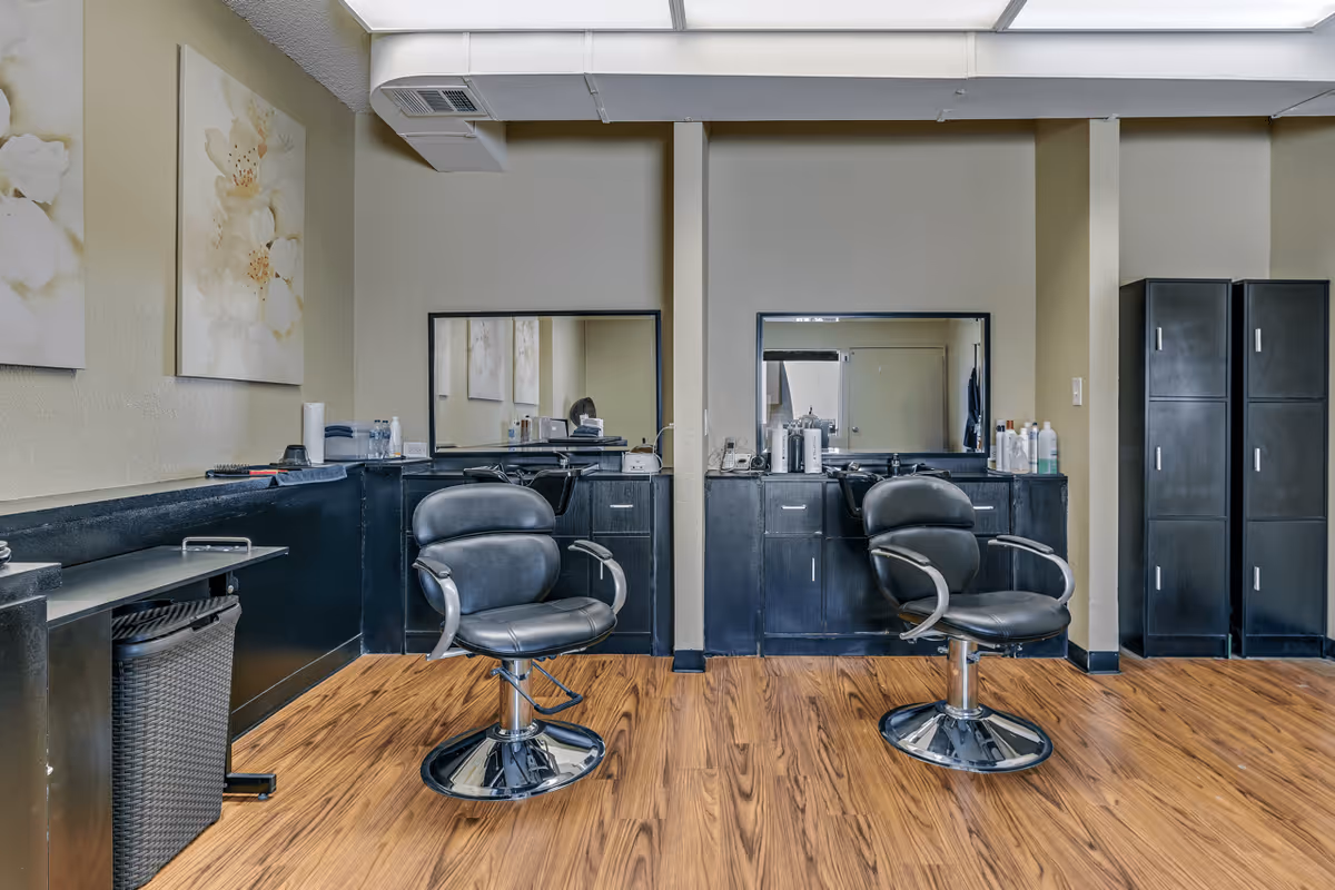 Interior view of a salon area with two black salon chairs in front of two mirrors mounted on a beige wall. The room has wooden flooring, black cabinets with various hair products on top, and two tall black storage cabinets on the right side. There are floral paintings on the left wall.