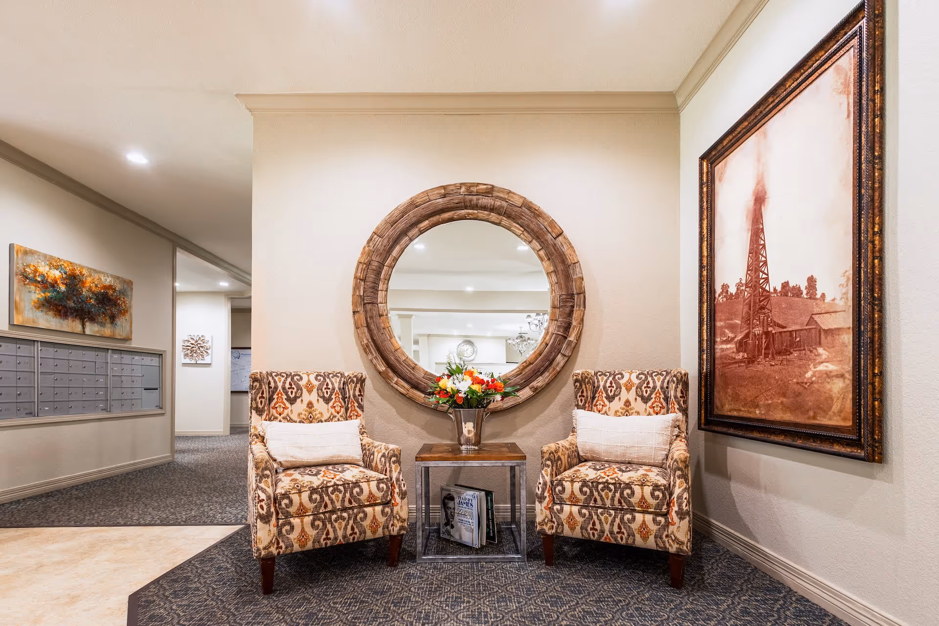 A cozy seating area in a senior living facility featuring two patterned armchairs with white cushions, a small wooden table with a vase of colorful flowers, and a large round wooden-framed mirror on the wall behind. To the right, there is a large framed sepia-toned photograph of an old oil derrick. On the left side, a hallway with mailboxes and artwork is visible.