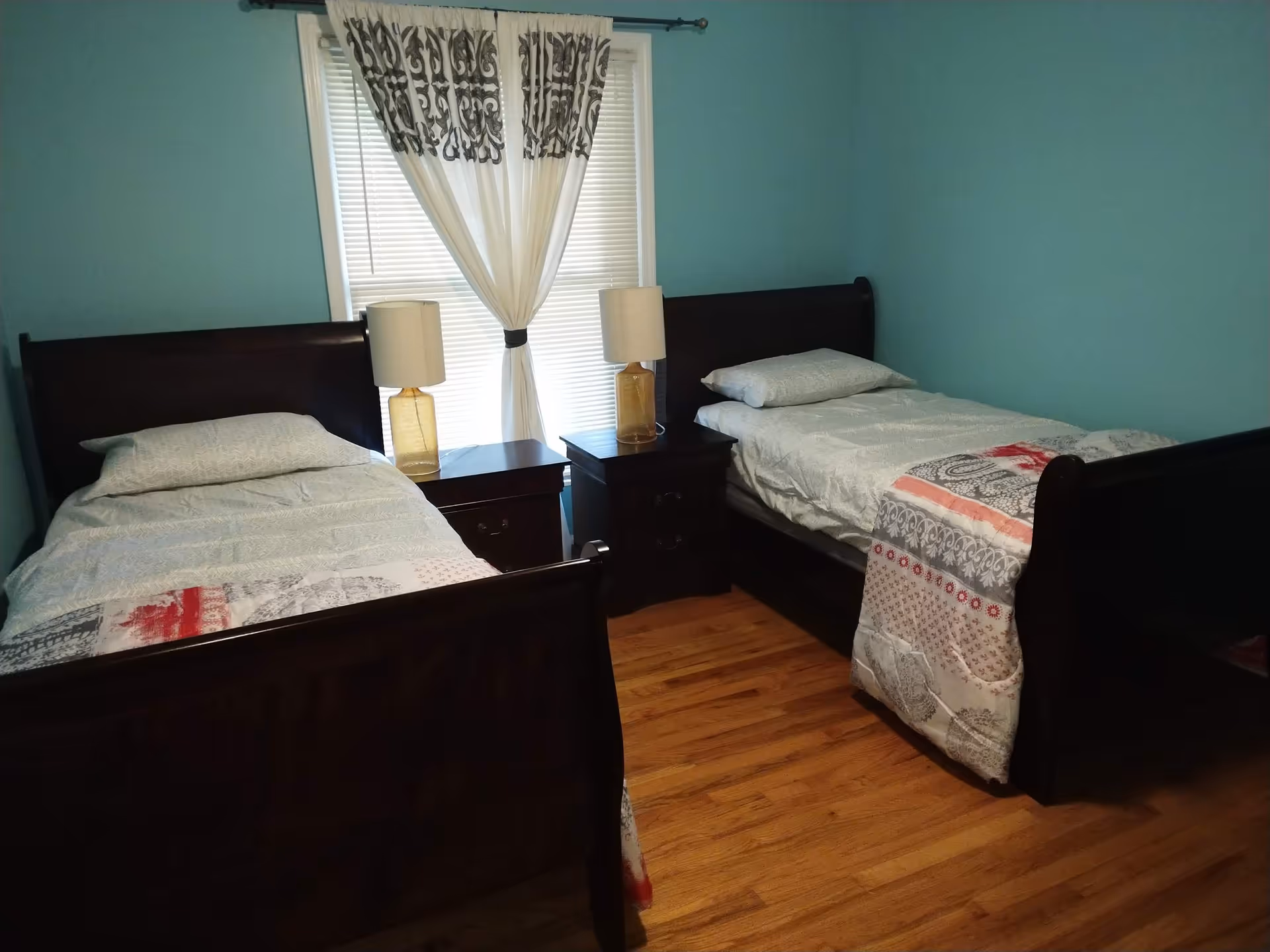 A bedroom with two single beds featuring dark wooden frames, each with white bedding and patterned quilts. Between the beds are two dark wooden nightstands, each with a lamp. A window with white blinds and a curtain with a decorative pattern is centered on the wall behind the nightstands. The walls are painted light blue and the floor is wooden.