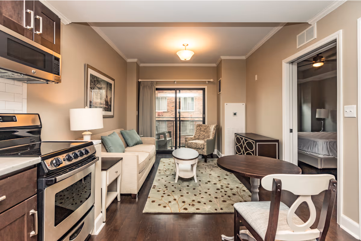 A cozy living area in an apartment at The Merion featuring a beige sofa with green cushions, a patterned armchair, a small oval coffee table on a decorative rug, and a round dining table with chairs. The kitchen area with a stove and microwave is visible on the left. A sliding glass door leads to a small balcony with outdoor chairs. A bedroom with a bed and lamp is visible through an open doorway on the right.