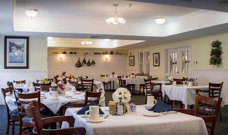 A dining room with multiple tables covered in white tablecloths, each set with plates, cups, silverware, and napkins. The room has wooden chairs, floral centerpieces on the tables, framed artwork on the walls, and decorative wall-mounted sailboats. The lighting includes ceiling fixtures and wall sconces, creating a warm and inviting atmosphere.