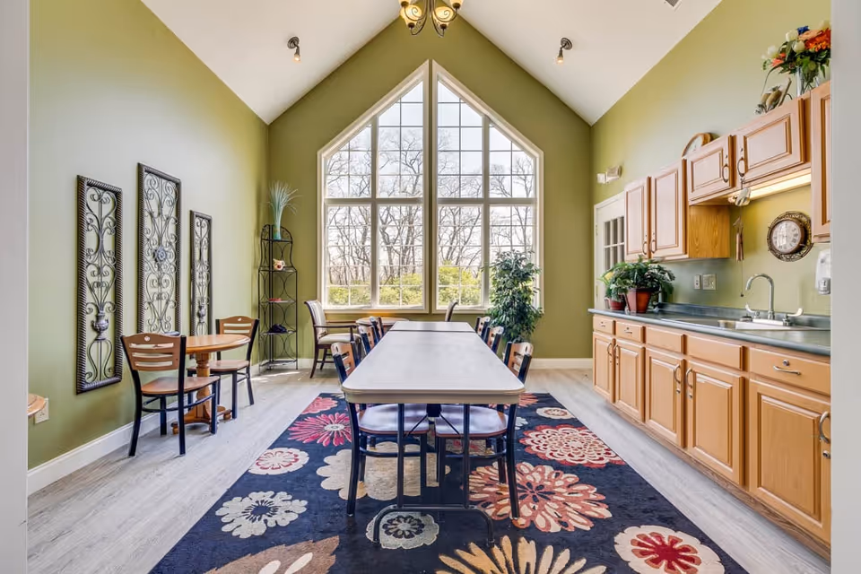 A bright dining room with a long rectangular table surrounded by chairs on a colorful floral rug. The room has high vaulted ceilings and a large window with multiple panes letting in natural light. On the right side, there is a kitchen counter with wooden cabinets, a sink, and some potted plants. On the left wall, there are three decorative wrought iron wall hangings and a small round table with two chairs. The walls are painted green.