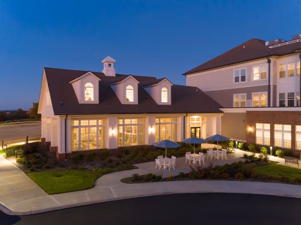 Evening exterior view of a senior living facility building with large windows lit from inside, outdoor patio area with tables, chairs, and umbrellas, surrounded by landscaped greenery and a curved driveway.