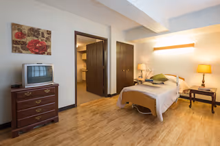 A senior living facility bedroom with a single bed covered in beige bedding and green pillows. There is a wooden nightstand with a lamp on the right side of the bed and another small table with a lamp and decorative items. A wooden dresser with an old-style TV sits against the left wall. The room has wooden flooring, a closet with double doors, and an open doorway leading to a bathroom area. A floral painting hangs on the wall above the TV.