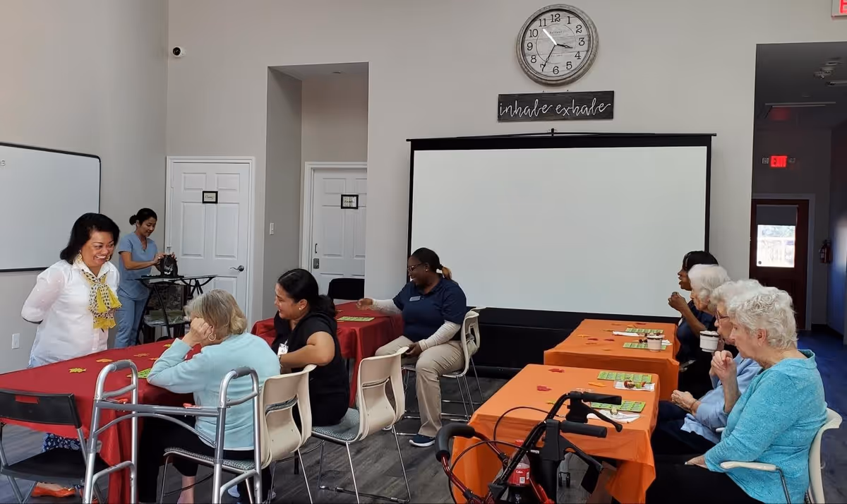 A group of elderly women and caregivers sitting around tables covered with red and orange tablecloths in a community room. Some are engaged in conversation, and there is a walker and a wheelchair visible. A large clock and a sign that reads 'inhale exhale' are on the wall above a blank projector screen.