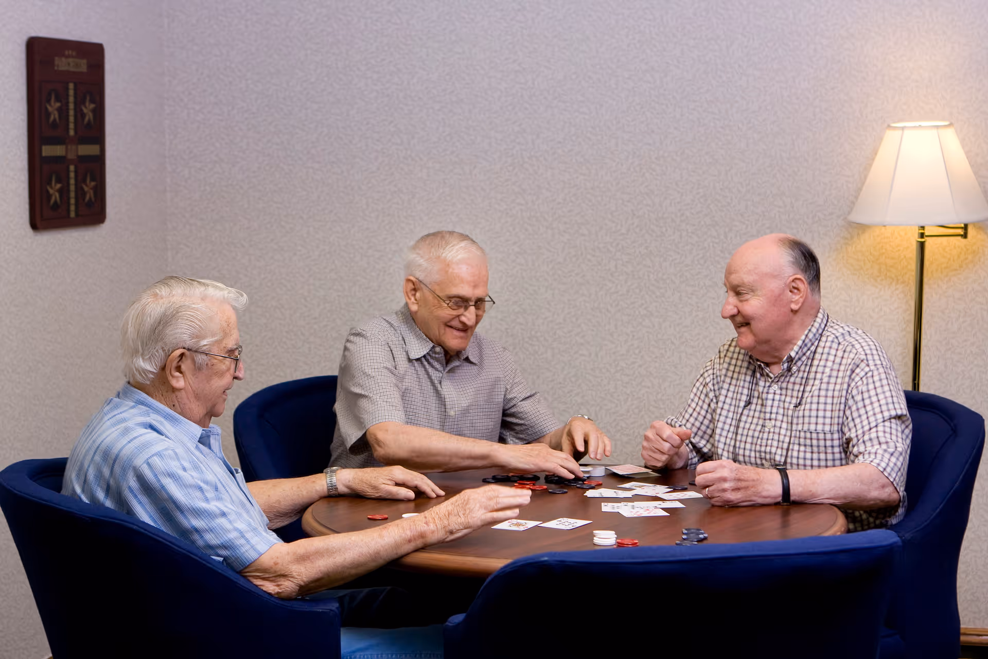 Three elderly men sitting around a round wooden table playing cards and poker chips in a cozy room with a floor lamp and wall decoration.