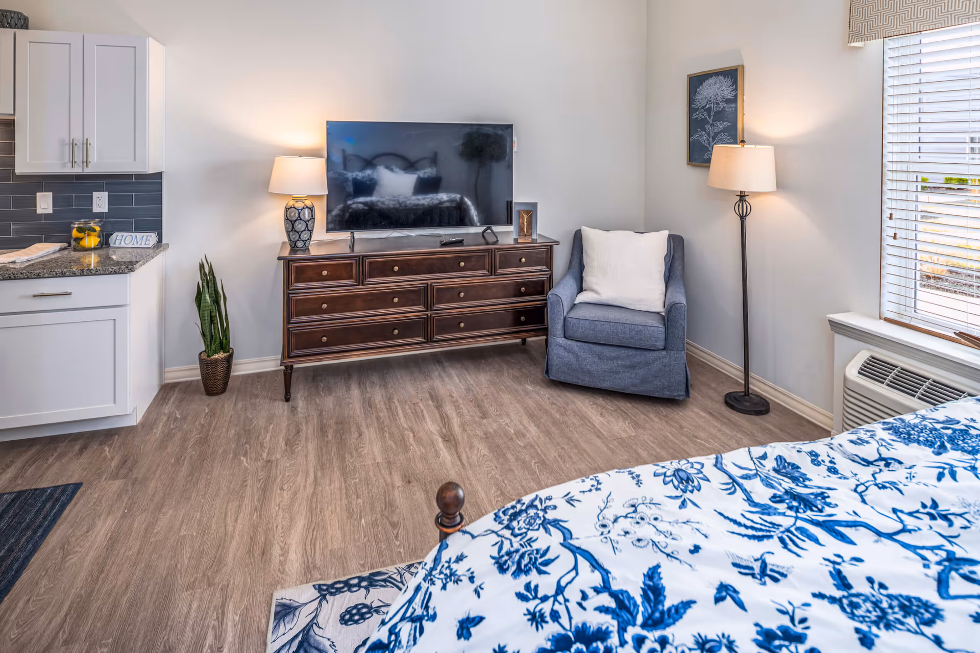 Studio apartment interior showing a bed corner in the foreground, a TV on a wooden dresser, a blue armchair, kitchenette cabinets, and a window with blinds.