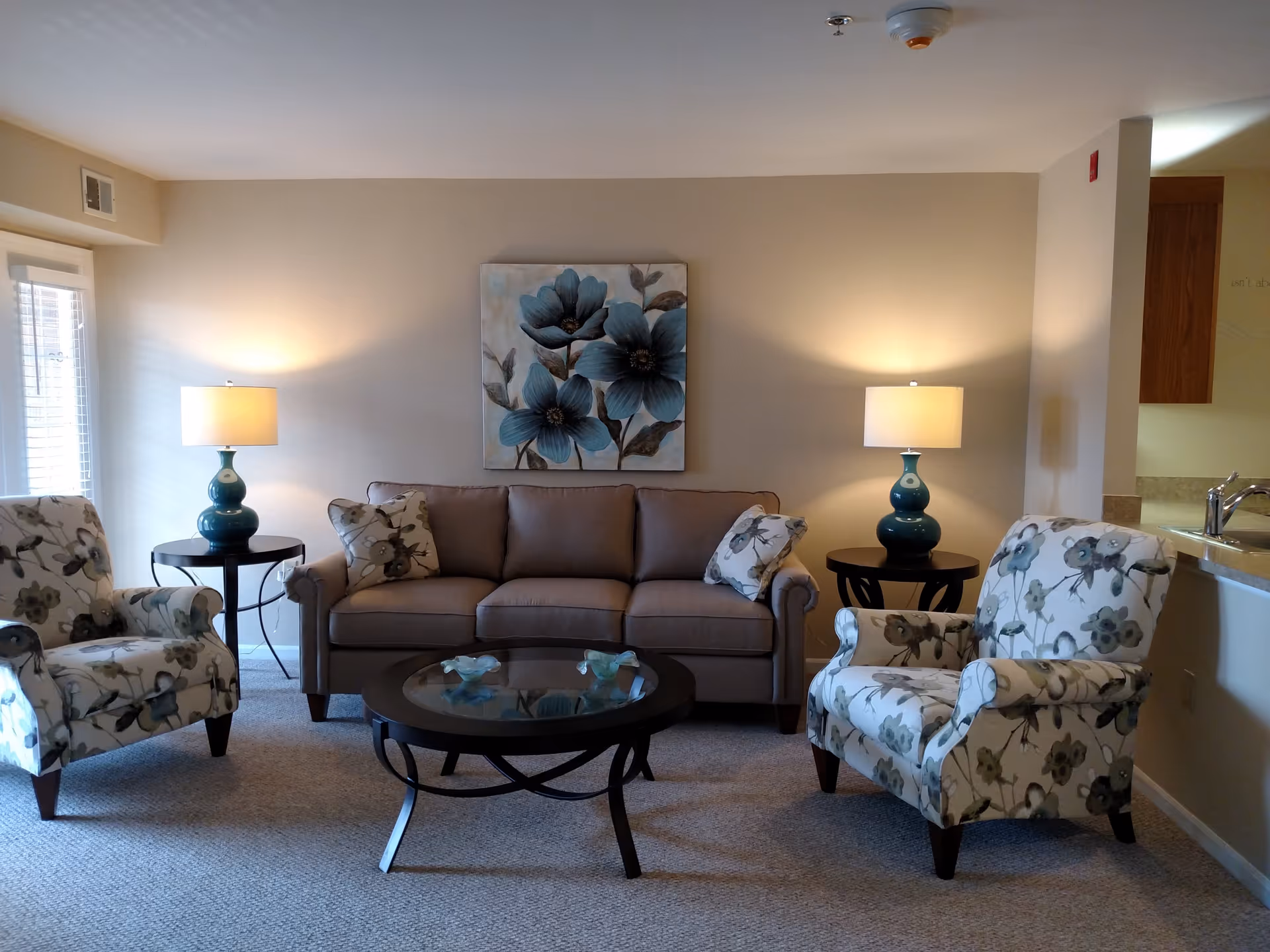 A cozy living room with a beige sofa adorned with floral pillows, flanked by two floral-patterned armchairs. Two matching teal lamps on round black side tables illuminate the room. A round glass coffee table sits in the center on a carpeted floor. A painting of large blue flowers hangs on the beige wall behind the sofa. The room has a warm and inviting atmosphere with a partial view of a kitchen counter and sink on the right.