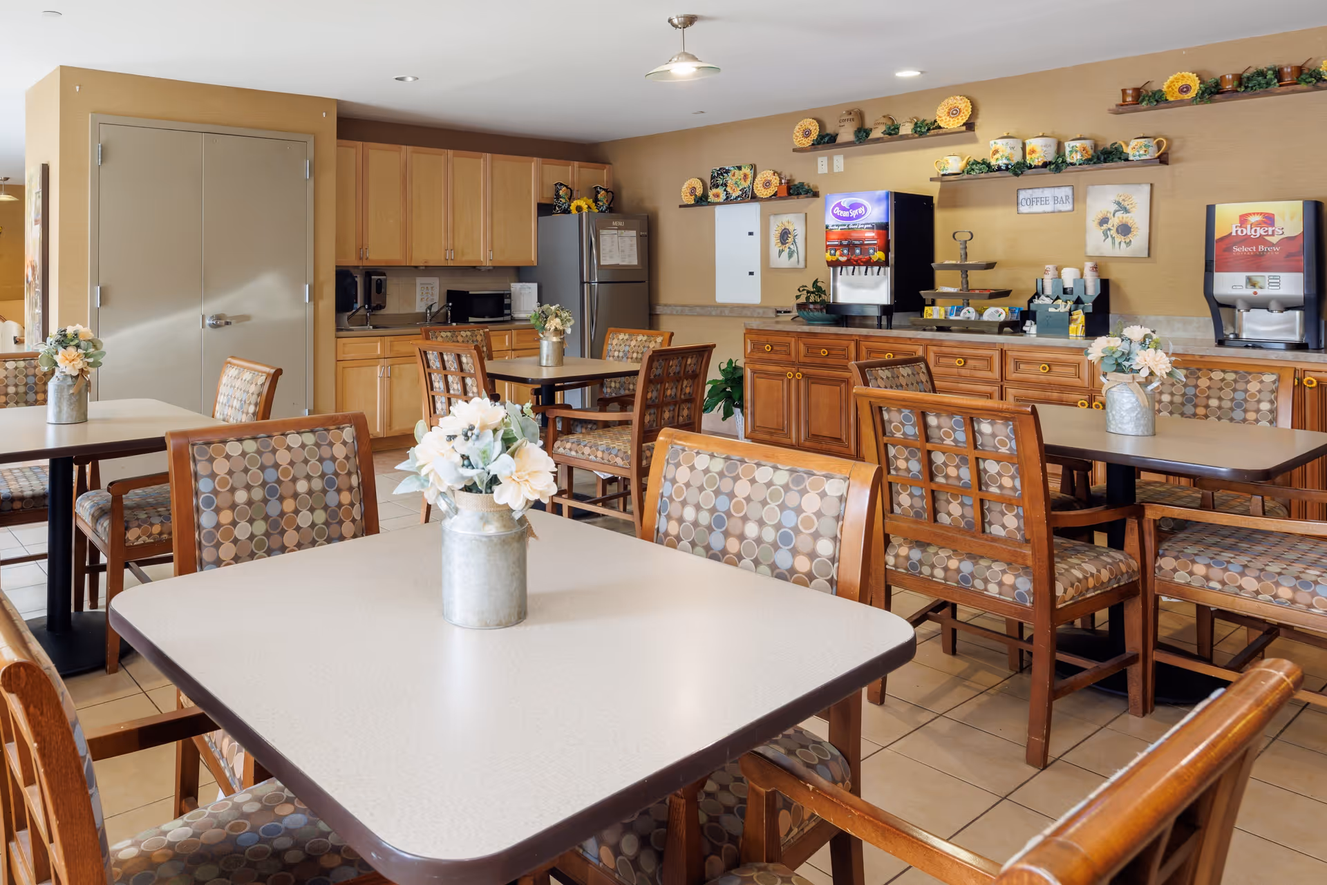 A cozy dining area with several tables and chairs featuring patterned upholstery. Each table has a small vase with white flowers. In the background, there is a kitchen area with wooden cabinets, a refrigerator, and a coffee bar with beverage dispensers and sunflower-themed decorations on the wall.