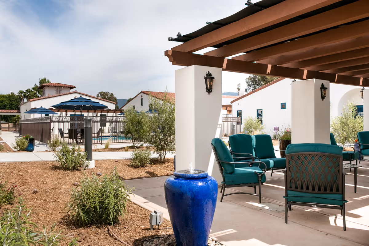 Outdoor seating area with teal cushioned chairs under a wooden pergola, a blue ceramic water fountain in the foreground, and white buildings with red tile roofs in the background. There are also blue umbrellas and a fenced swimming pool visible.