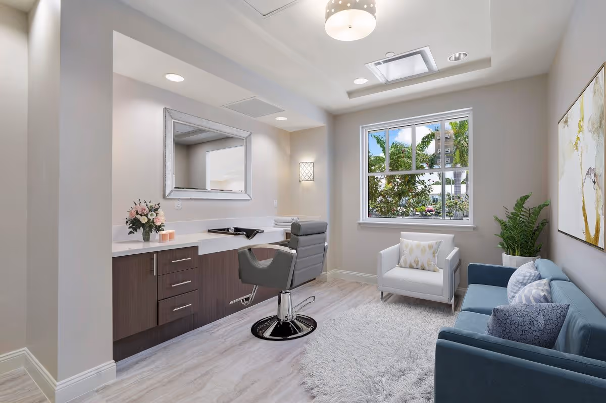 Well-lit lounge with a salon-style chair and counter under a mirror, a sofa and armchair, and a window overlooking palm trees.