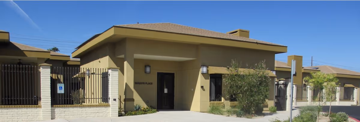 Exterior front view of a single-story senior living facility building named Sarah's Place, featuring beige walls, a covered entrance, and surrounding landscaping with bushes and small trees under a clear blue sky.