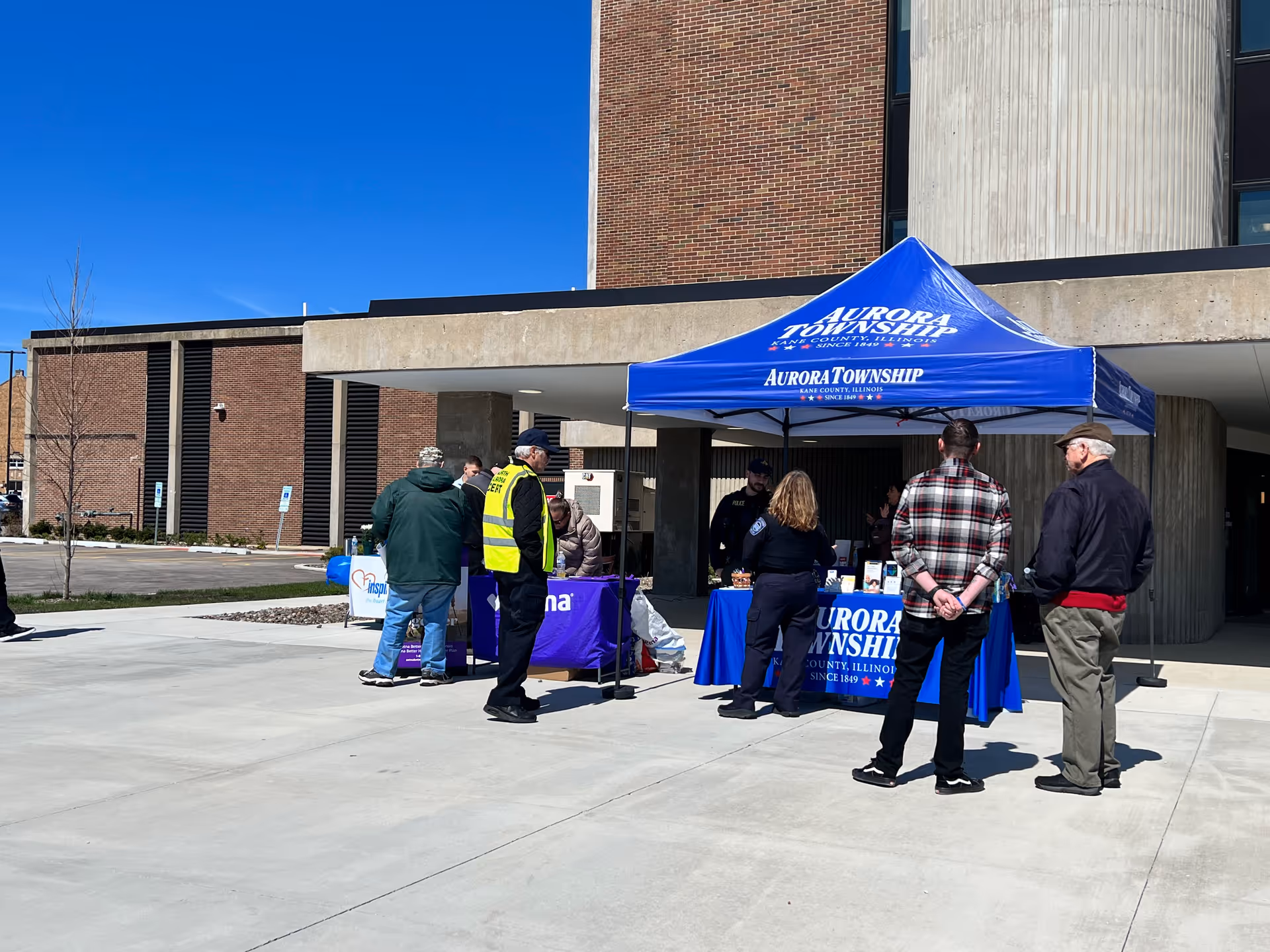 Several people gathered around a blue "Aurora Township" canopy and information table outside a brick building under a clear blue sky.