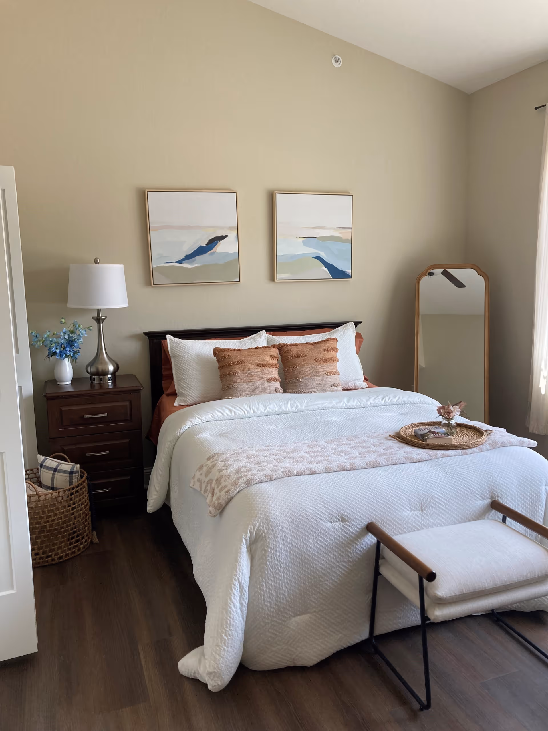 A neatly made bed with white bedding and two brown decorative pillows in a bedroom. There is a wooden nightstand with a silver lamp and a vase with blue flowers on the left side of the bed. Two abstract paintings hang on the beige wall above the bed. A wicker basket with pillows is on the floor next to the nightstand. A full-length mirror with a wooden frame stands against the right wall near a window with light curtains. A small bench with a cushion is placed at the foot of the bed.