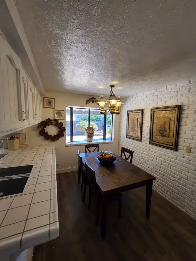 A cozy dining area with a wooden table and four chairs, a bowl of fruit on the table, white brick wall with two framed pictures, a window overlooking a garden, and a chandelier with three lights hanging from the ceiling. The adjacent kitchen counter has a tiled surface and white cabinets above.