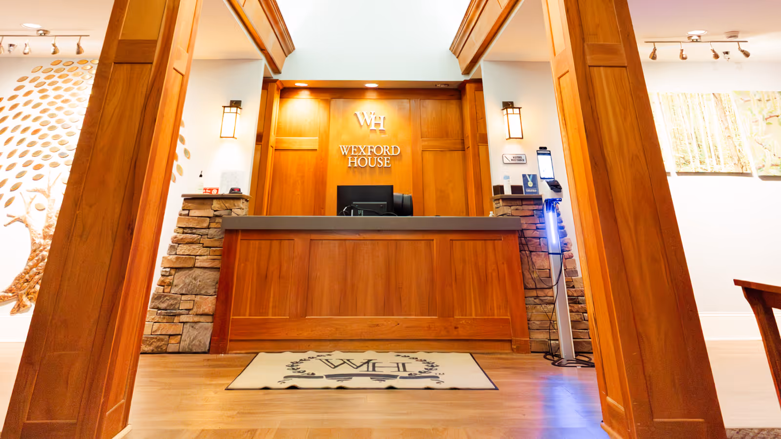 Reception desk area at Wexford House with wooden paneling and stone accents. The Wexford House logo is displayed on the wall behind the desk. There is a mat on the floor with the initials WH, and a hand sanitizer dispenser is visible to the right of the desk. The area is well-lit with wall sconces and overhead lights.