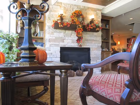 Cozy living room area with a stone fireplace decorated with autumn-themed wreath and garland. There are two upholstered chairs with wooden frames and a wooden side table with a decorative lamp and a small pumpkin on it. The room has warm lighting and a comfortable, inviting atmosphere.