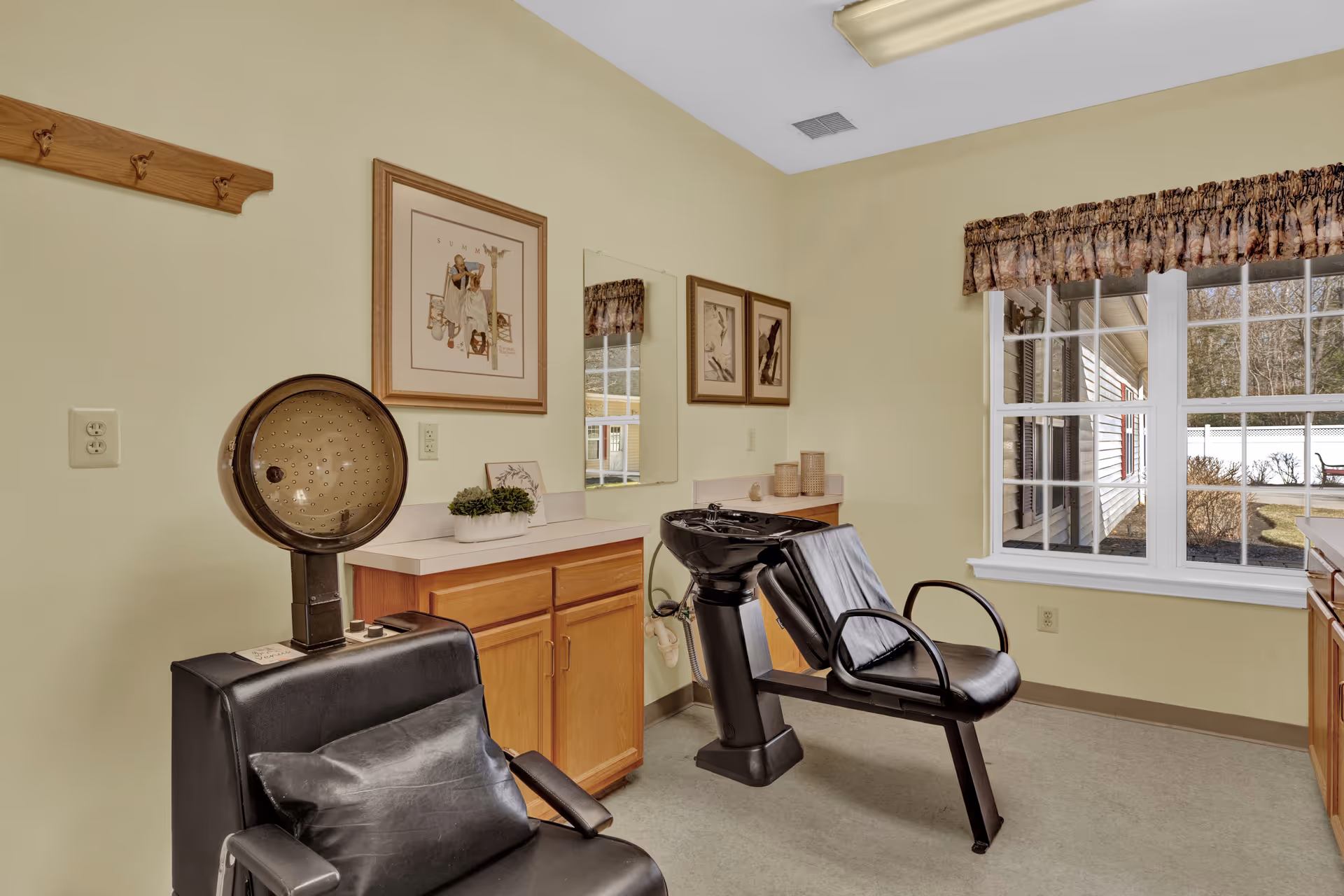 Small salon room with a hooded hair dryer chair, shampoo sink and styling chair, wooden cabinets, framed art, and a large window.