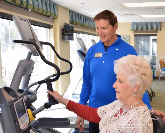 An elderly woman using an elliptical exercise machine in a fitness room while a male staff member in a blue jacket stands beside her, smiling and providing assistance. The room has large windows with blue valances and exercise equipment visible in the background.