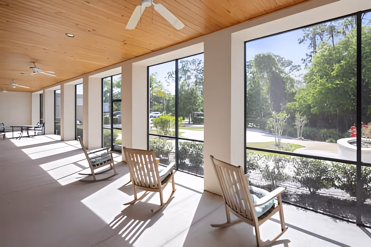 A bright, screened-in porch area with wooden rocking chairs and a wooden ceiling with ceiling fans. Large windows provide a view of greenery and a driveway outside.