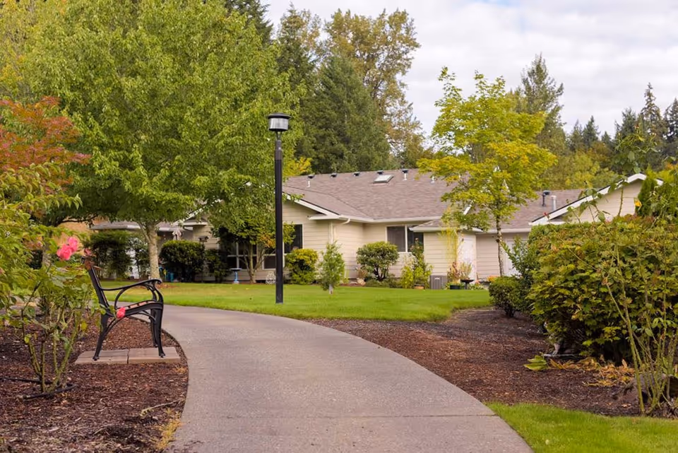 A paved walkway curves through a landscaped garden area with green grass, bushes, and trees. A black metal bench is positioned on the left side of the path near some pink flowers. In the background, there is a single-story beige building with a gray roof, surrounded by more greenery under a partly cloudy sky.