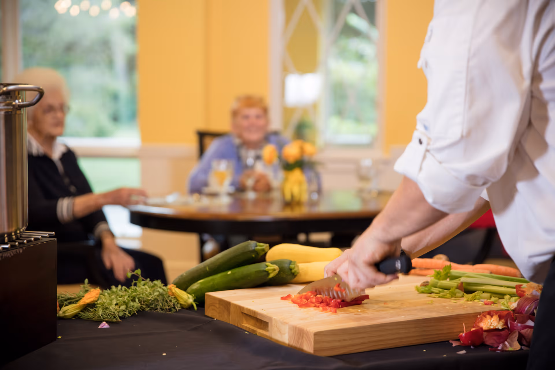 A person chopping red bell peppers on a wooden cutting board with various vegetables like zucchini, carrots, and celery nearby. In the background, two elderly women are seated at a dining table with glasses and a vase of yellow flowers, in a room with yellow walls and large windows.