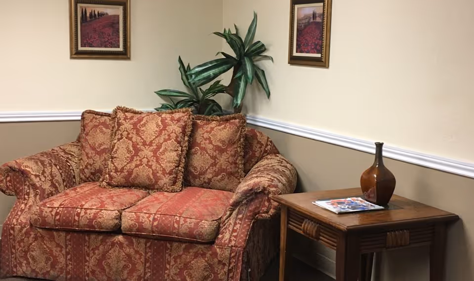 A cozy corner of a room featuring a patterned red and gold loveseat with three matching cushions. Behind the loveseat is a green leafy plant. On the beige and light brown walls, there are two framed landscape paintings. Next to the loveseat is a wooden side table with a brown vase and some magazines on top.