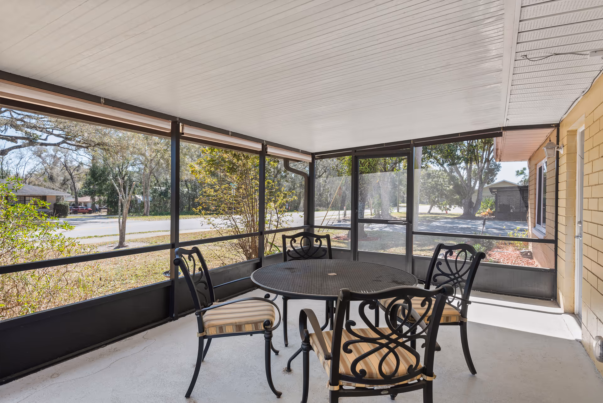A screened-in porch area with a round metal table and four metal chairs with striped cushions. The porch overlooks a yard with trees and a street in the background. The porch has a white ceiling and a concrete floor.