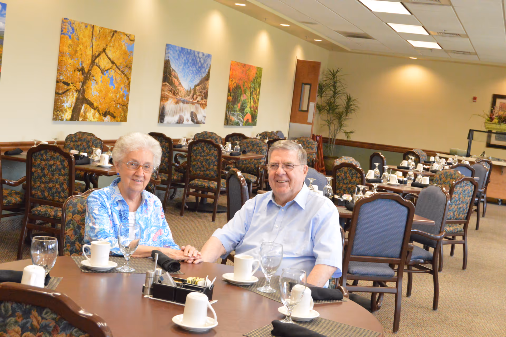 Two elderly people seated at a table in a dining room with set tables and landscape artwork on the walls.
