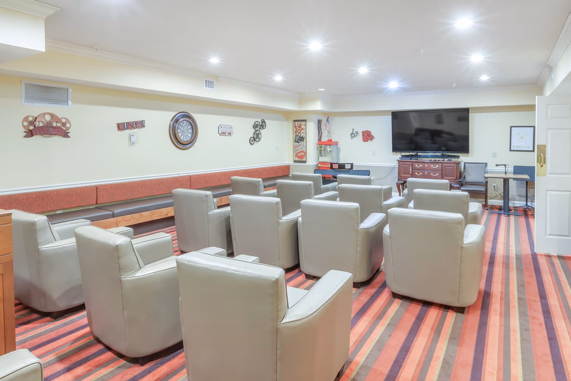A lounge/media room with rows of beige armchairs facing a wall-mounted television, bench seating along the wall and a striped carpet.