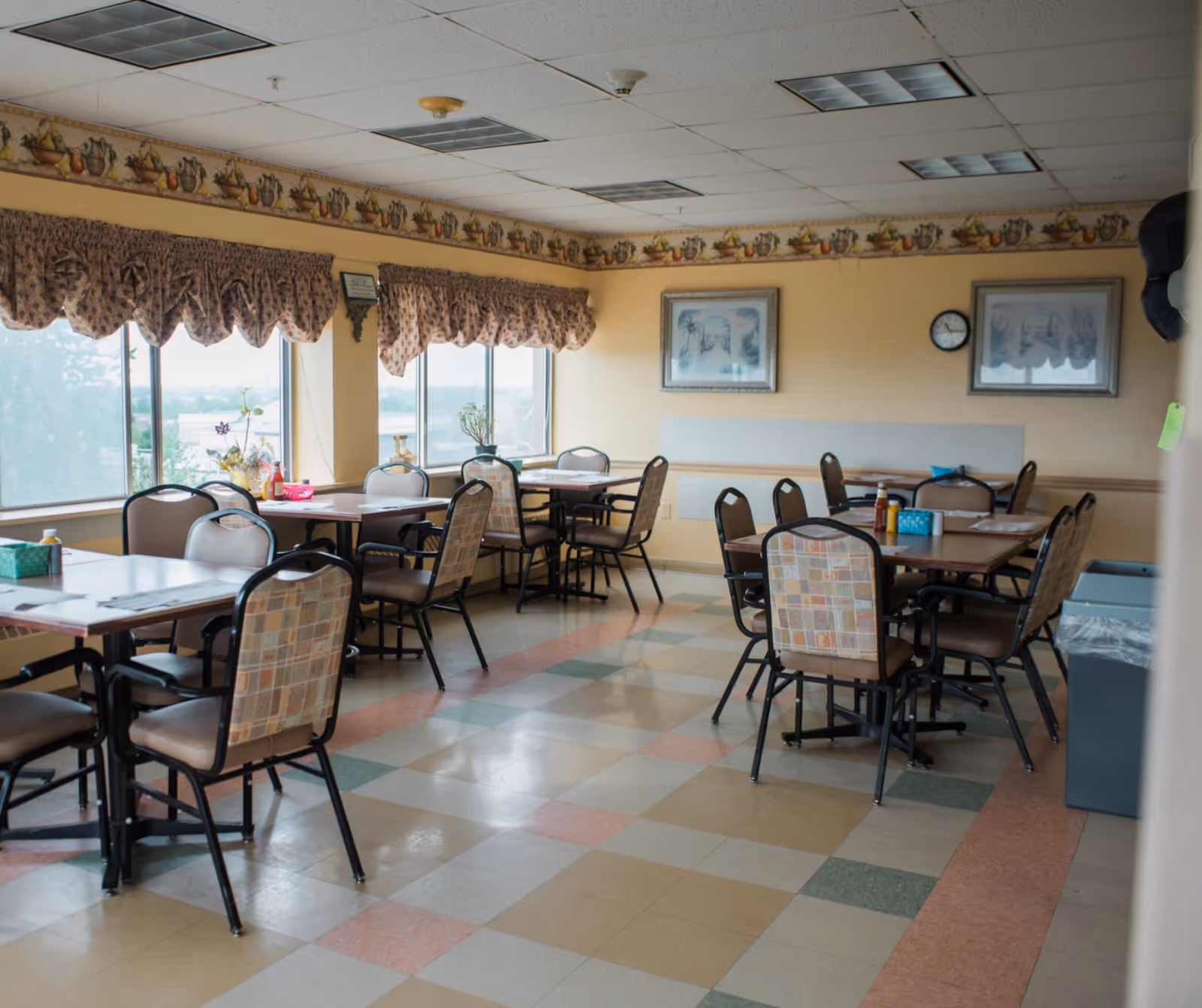 A dining room in Mary Agnes Manor with several tables and chairs arranged neatly. The room has large windows with patterned valances, framed artwork on the walls, and a clock. The floor is tiled with a multicolored pattern, and there are condiments and napkin holders on the tables.