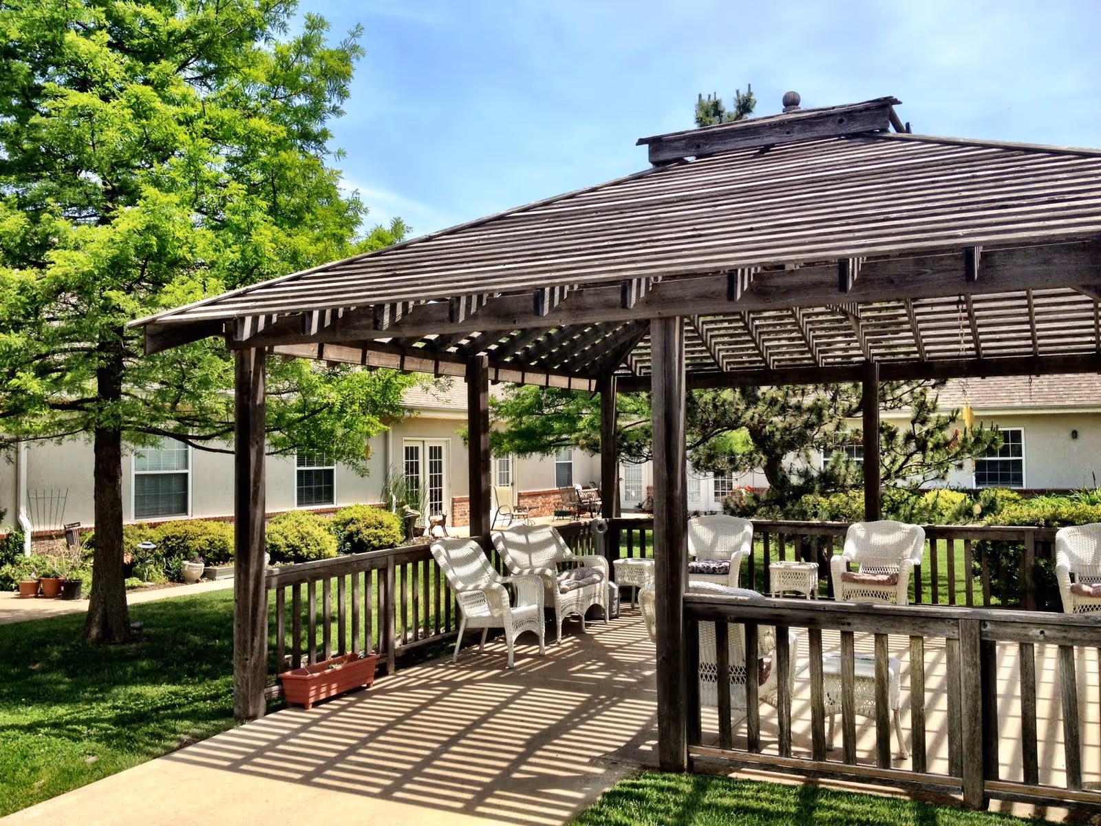 Wooden pergola over a patio with white wicker chairs and tables in a grassy courtyard outside a single-story assisted living building.