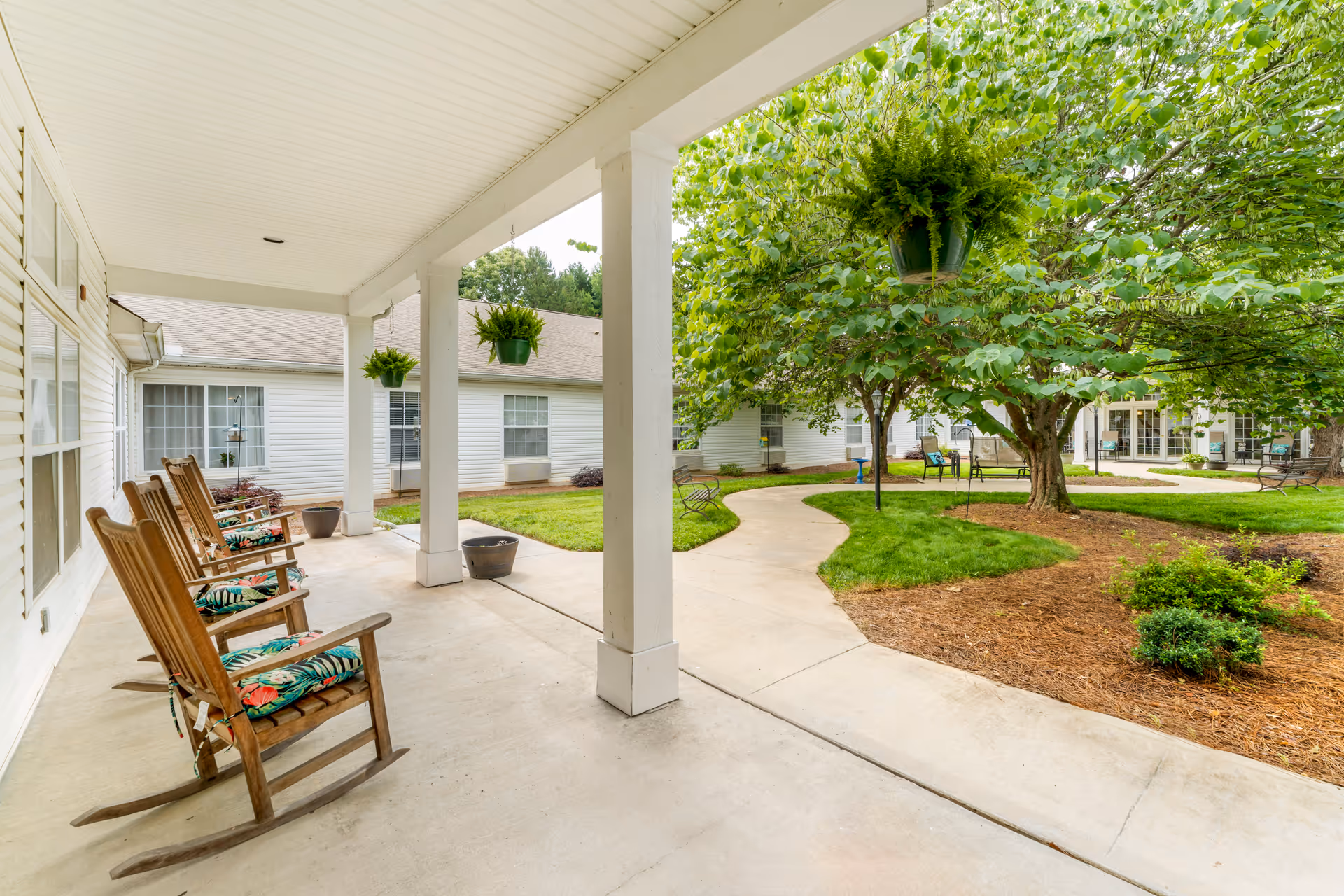 Covered porch with wooden rocking chairs and hanging plants overlooking a landscaped courtyard with walkways and trees.