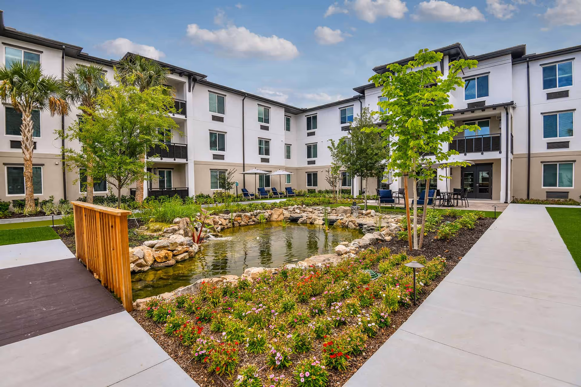Outdoor courtyard area of a senior living facility featuring a small pond surrounded by rocks and colorful flowers, with trees and shrubs planted around. The three-story building with multiple windows and balconies surrounds the courtyard. There are lounge chairs with umbrellas and a seating area with tables and chairs on the paved walkway.