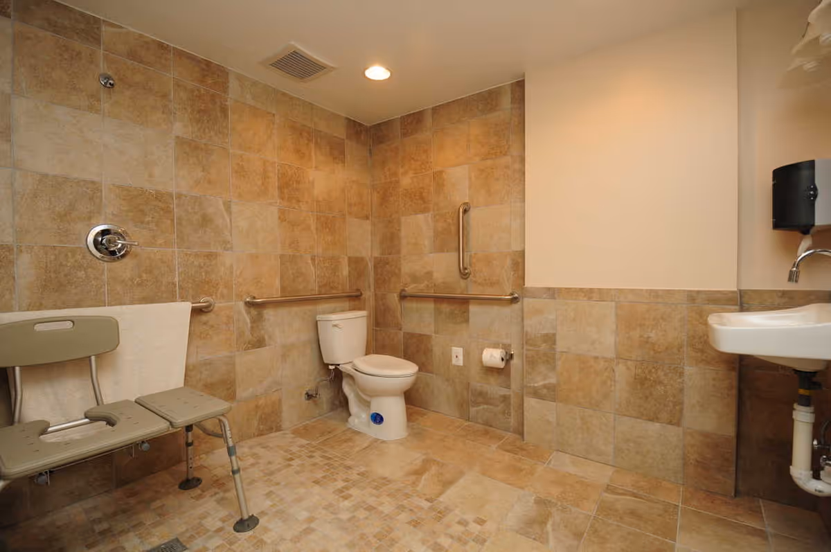 Accessible bathroom with beige tiled walls and floor, featuring a toilet with grab bars on adjacent walls, a wall-mounted sink with a soap dispenser above it, and a shower chair with a towel draped over it.