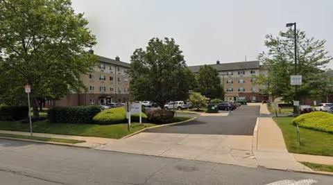 Wide view of Gloria Dei Estates senior living campus with a driveway, parking, and green landscaping.