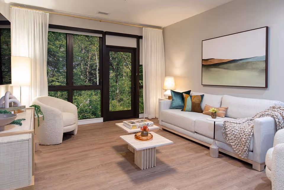 Contemporary living room with a light sofa, armchair, coffee tables, lamps, and large windows/door overlooking trees.