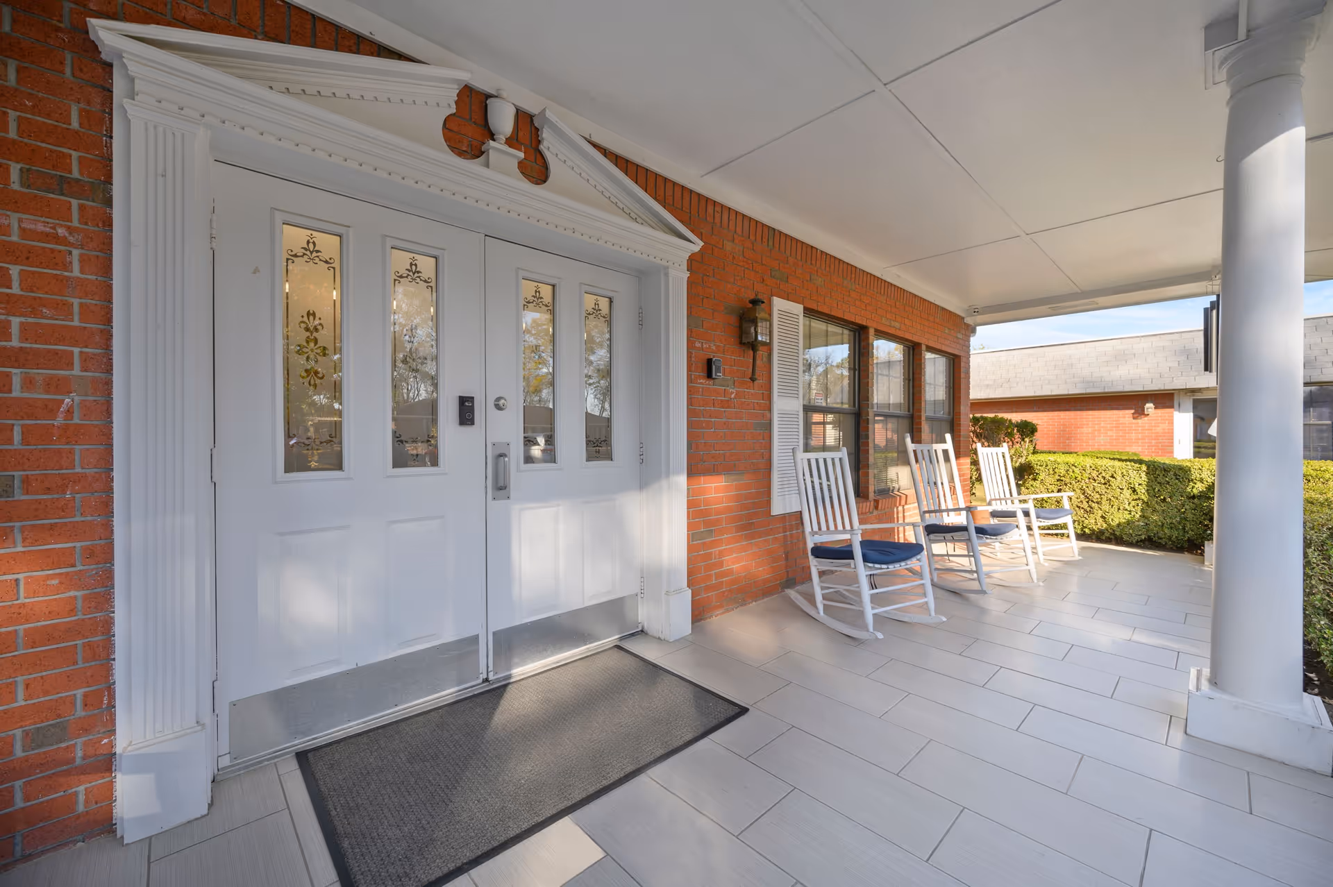 Covered front porch entrance with white double doors, rocking chairs, and tiled walkway at a senior living facility.
