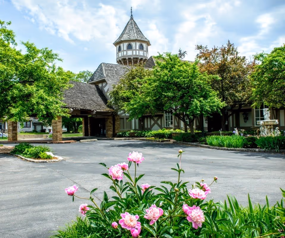 The front exterior of The Grand Regency facility featuring a Tudor-style building with a pointed tower, surrounded by lush green trees and blooming pink flowers in the foreground under a partly cloudy sky.