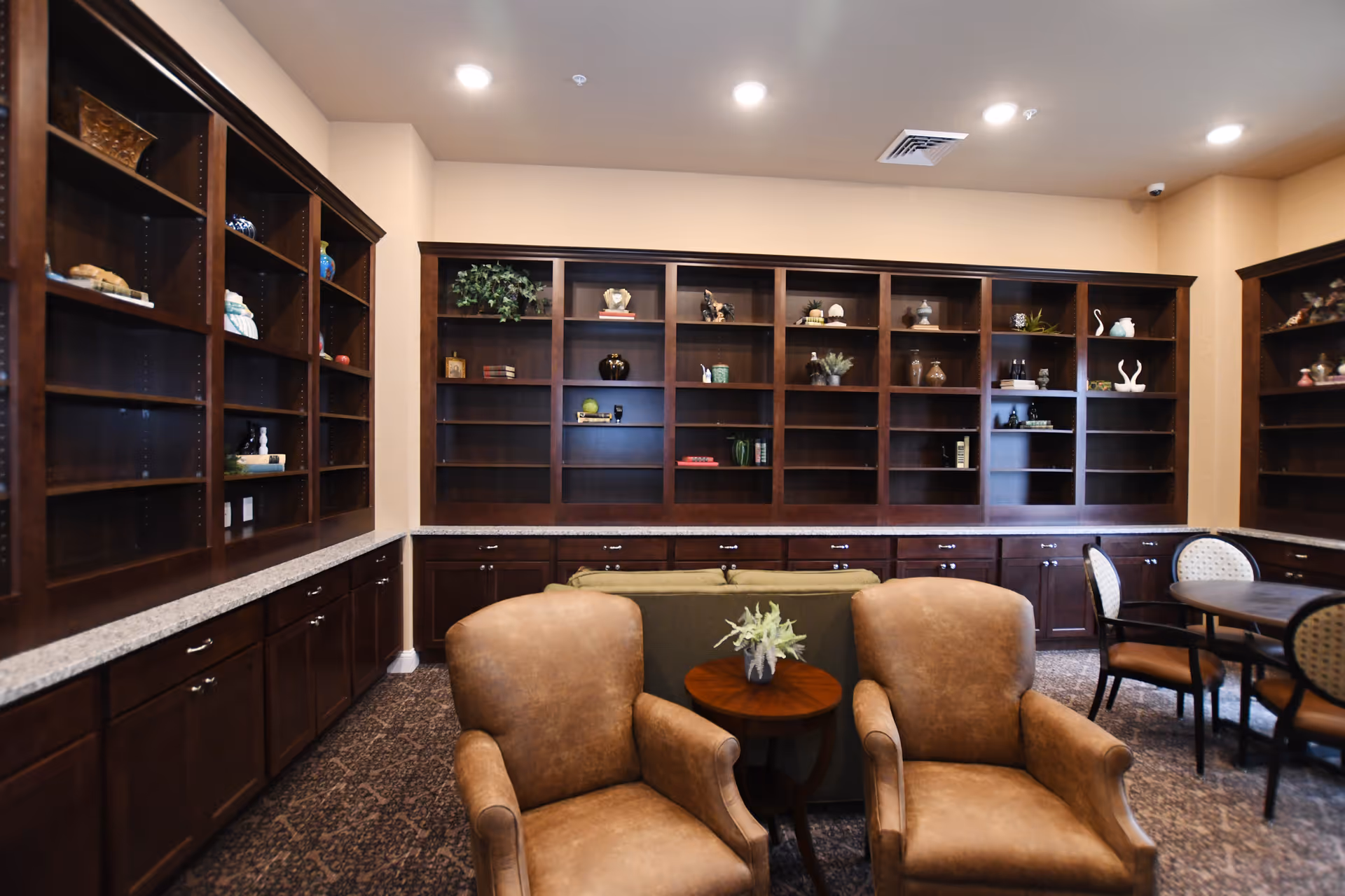A cozy common room with large built-in wooden bookshelves, upholstered armchairs, a small table and a seating area.
