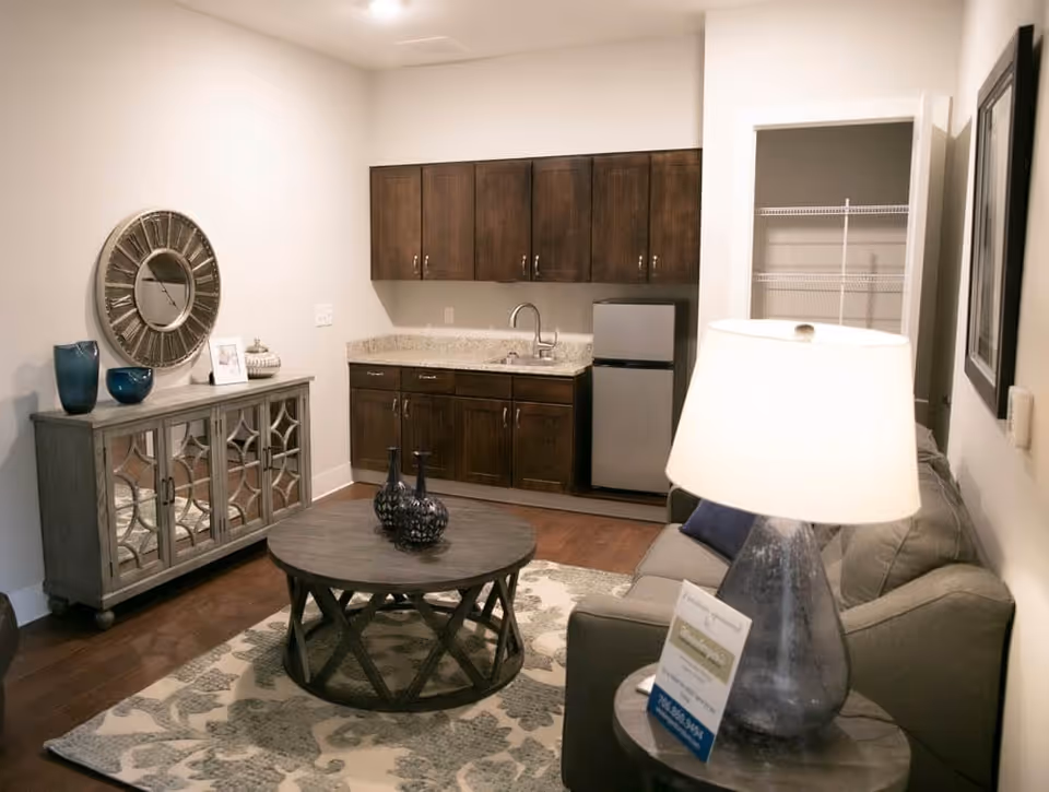 A cozy living area with a gray sofa, a round wooden coffee table with decorative vases, a side table with a lamp and informational card, a mirrored cabinet with decorative items and a large wall clock above it. In the background, there is a small kitchenette with dark wooden cabinets, a granite countertop, a sink, and a mini refrigerator. An open pantry with wire shelving is visible to the right of the kitchenette.