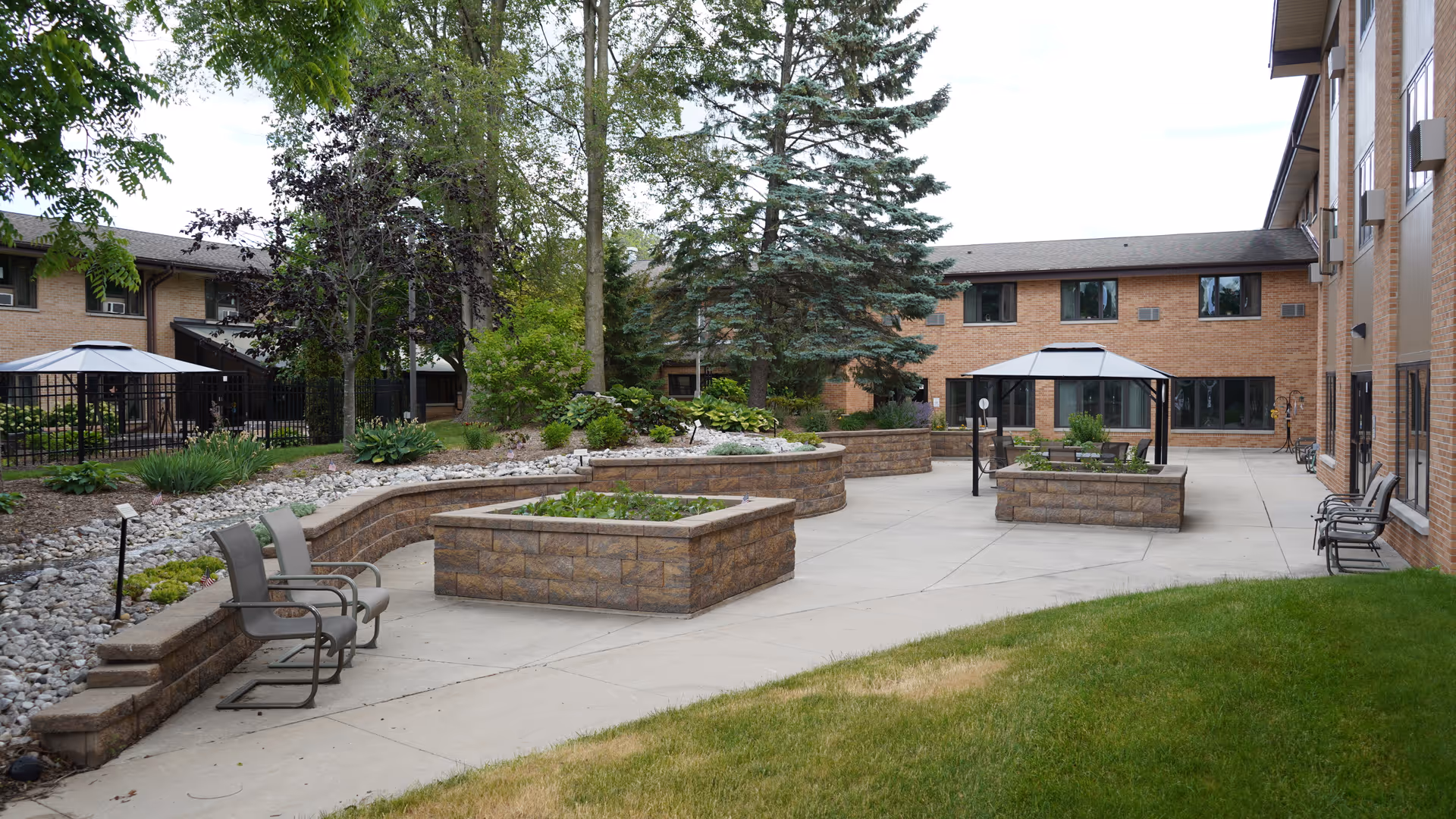 Outdoor courtyard area at Pine Haven Christian Communities Giddings Avenue Campus featuring raised garden beds, several chairs, two gazebos, trees, and surrounding brick buildings.