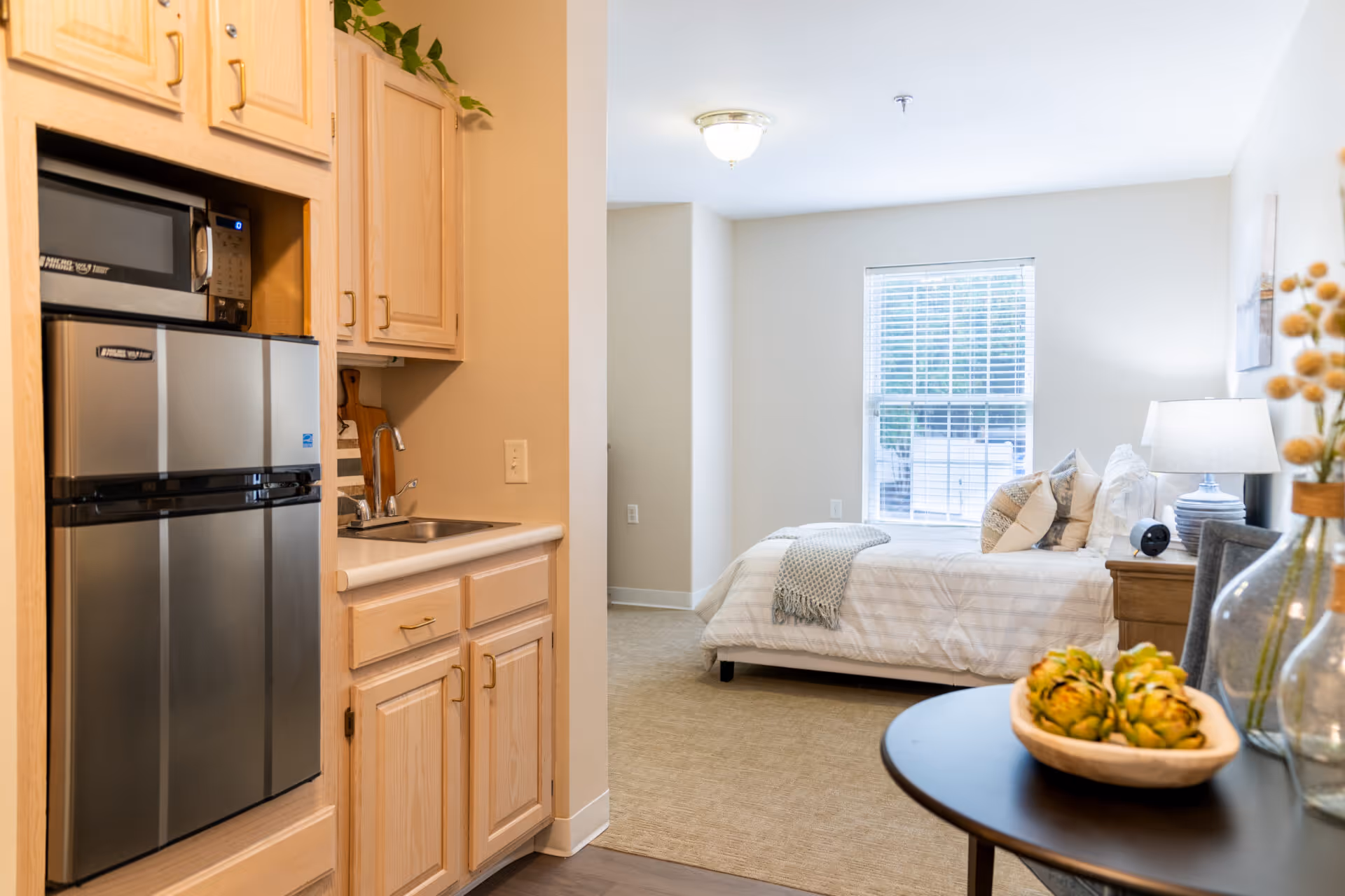 A bright and cozy studio apartment featuring a small kitchenette with a microwave, mini refrigerator, and sink on the left. The sleeping area has a neatly made bed with pillows and a throw blanket near a window with blinds. A bedside table with a lamp and decorative items is next to the bed. In the foreground, a round table holds a decorative bowl with artichokes and glass vases with dried flowers.