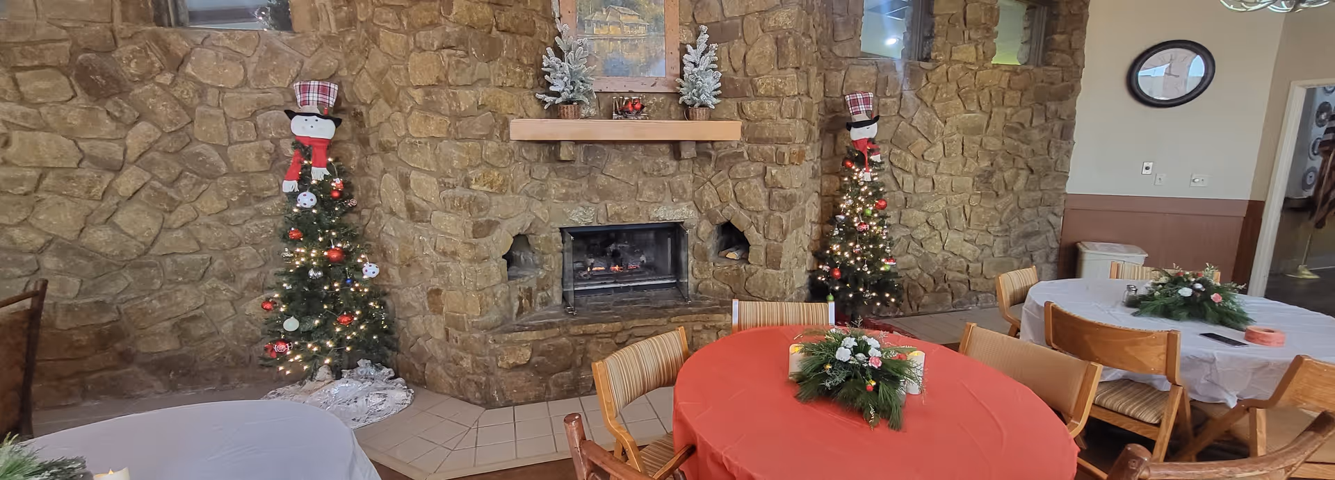 A cozy interior room with stone walls and a stone fireplace in the center. Two small decorated Christmas trees with snowman toppers flank the fireplace. Several round tables covered with red and white tablecloths are arranged around the room, each with a festive centerpiece. Wooden chairs surround the tables, and a framed picture hangs above the fireplace.